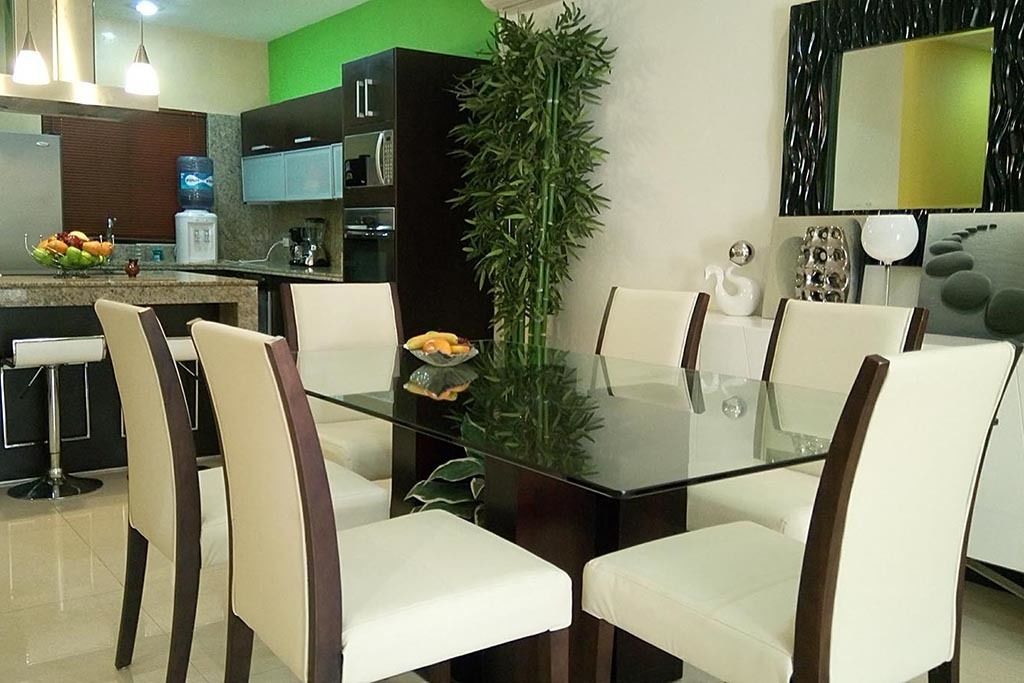 A modern dining area with a glass table surrounded by white chairs, a kitchen in the background, and a large green plant.