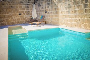 Una piscina con dos sillones de descanso y una tumbona pequeña, rodeada de muros de piedra.