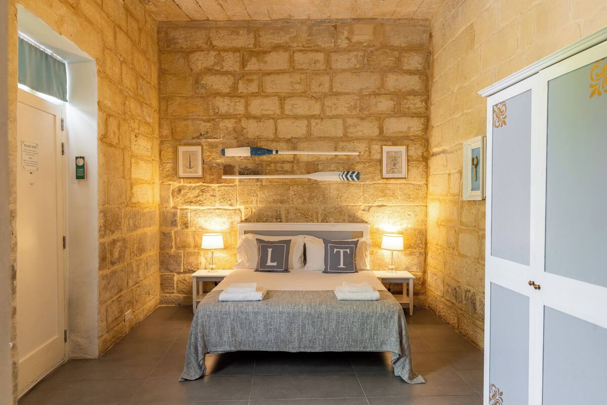 A bedroom with a stone wall, a bed with two pillows, a nightstand, and a shelf with decorative items.