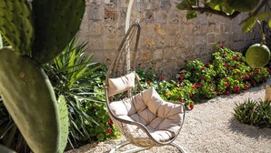 A wicker hanging chair with a cushioned seat in a garden setting with a stone wall and various plants. - Lemon Tree Relais by CX Collection (Zabbar)