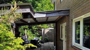 A covered patio area with a table and chairs, surrounded by greenery and a wooden fence.