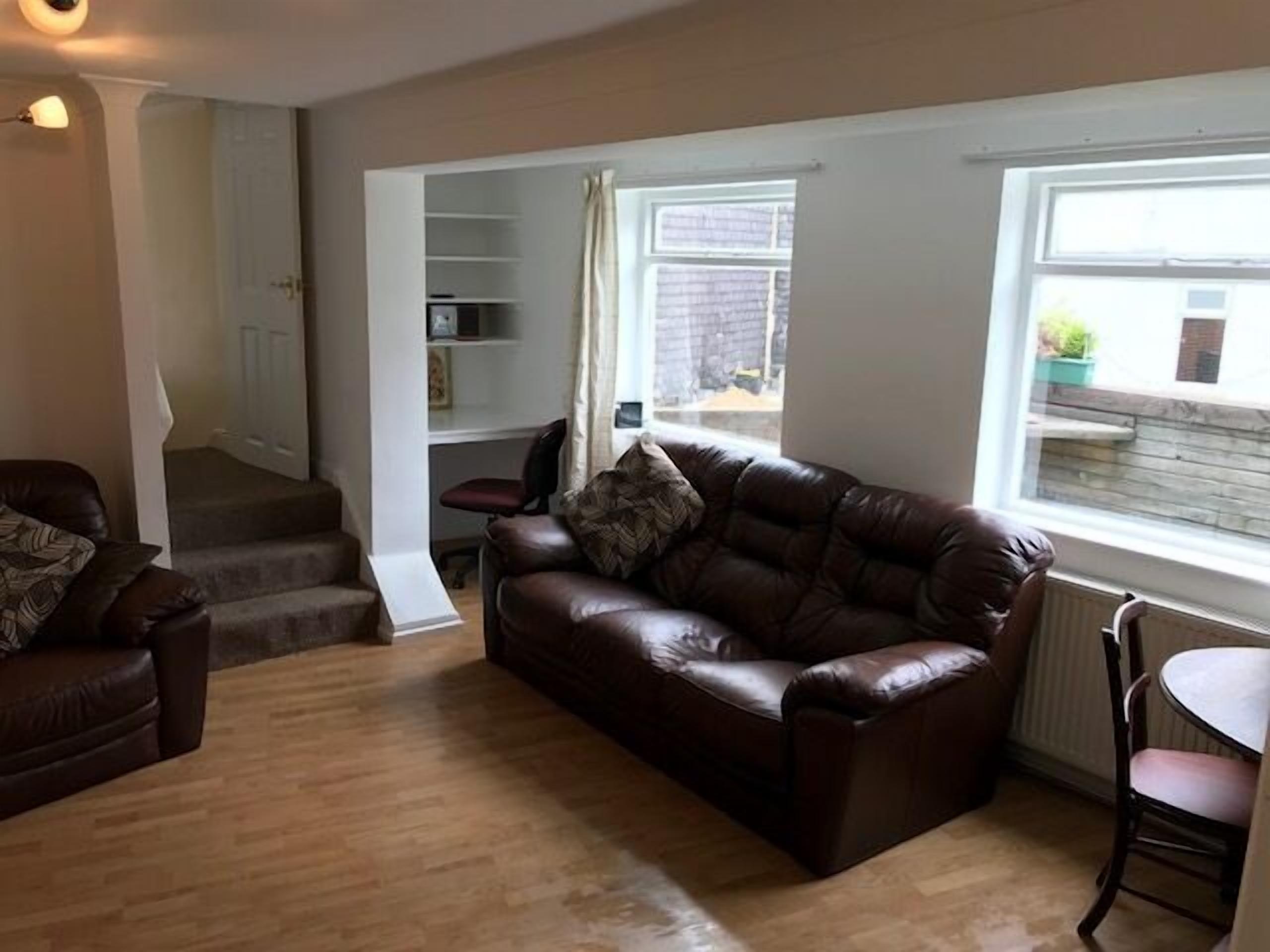 A living room with a brown leather sofa, a wooden floor, a dining table with a chair, and a staircase leading to a second floor.