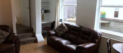 A living room with a brown leather sofa, a wooden floor, a dining table with a chair, and a staircase leading to a second floor.
