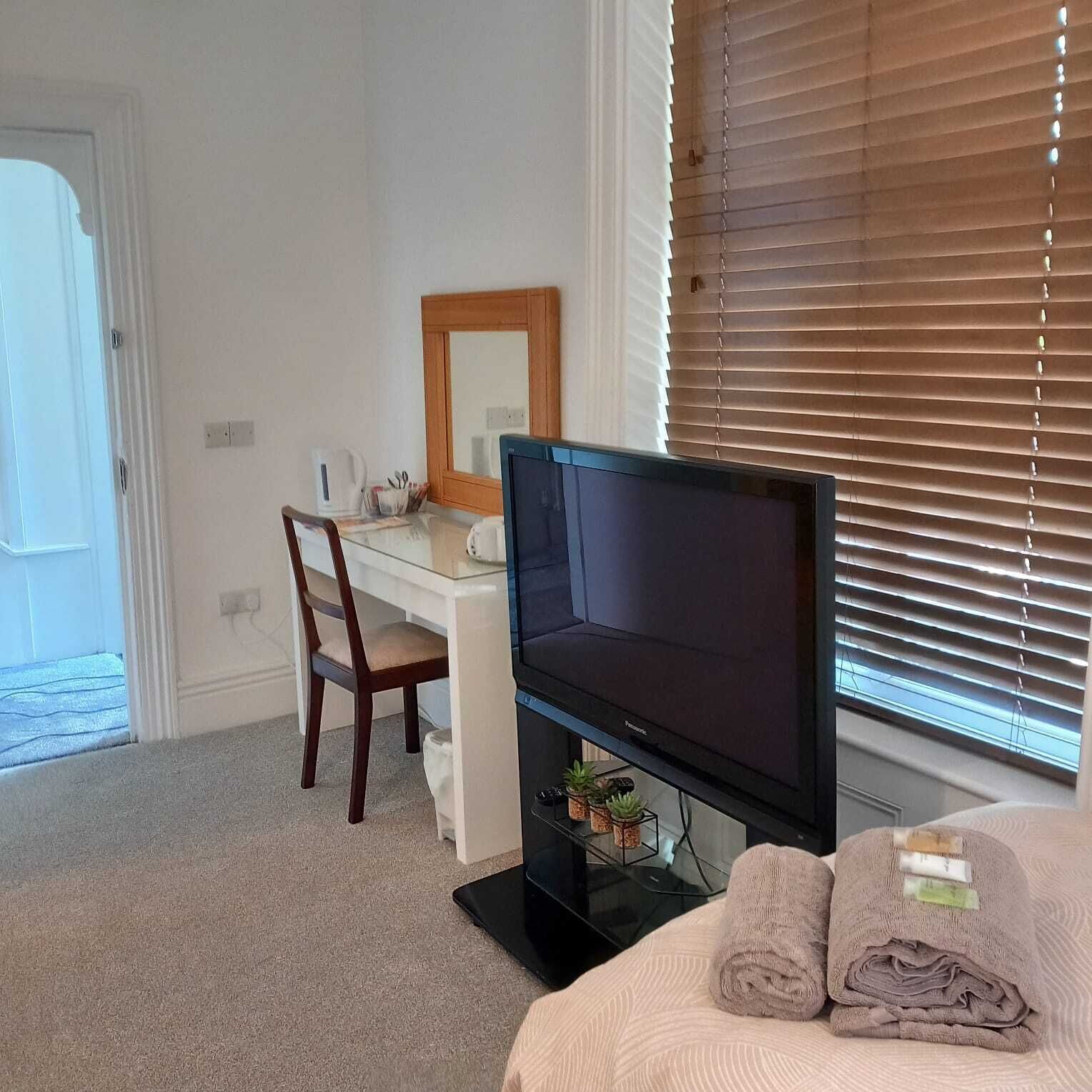 A bedroom with a bed, a desk, a television, and a window with blinds.