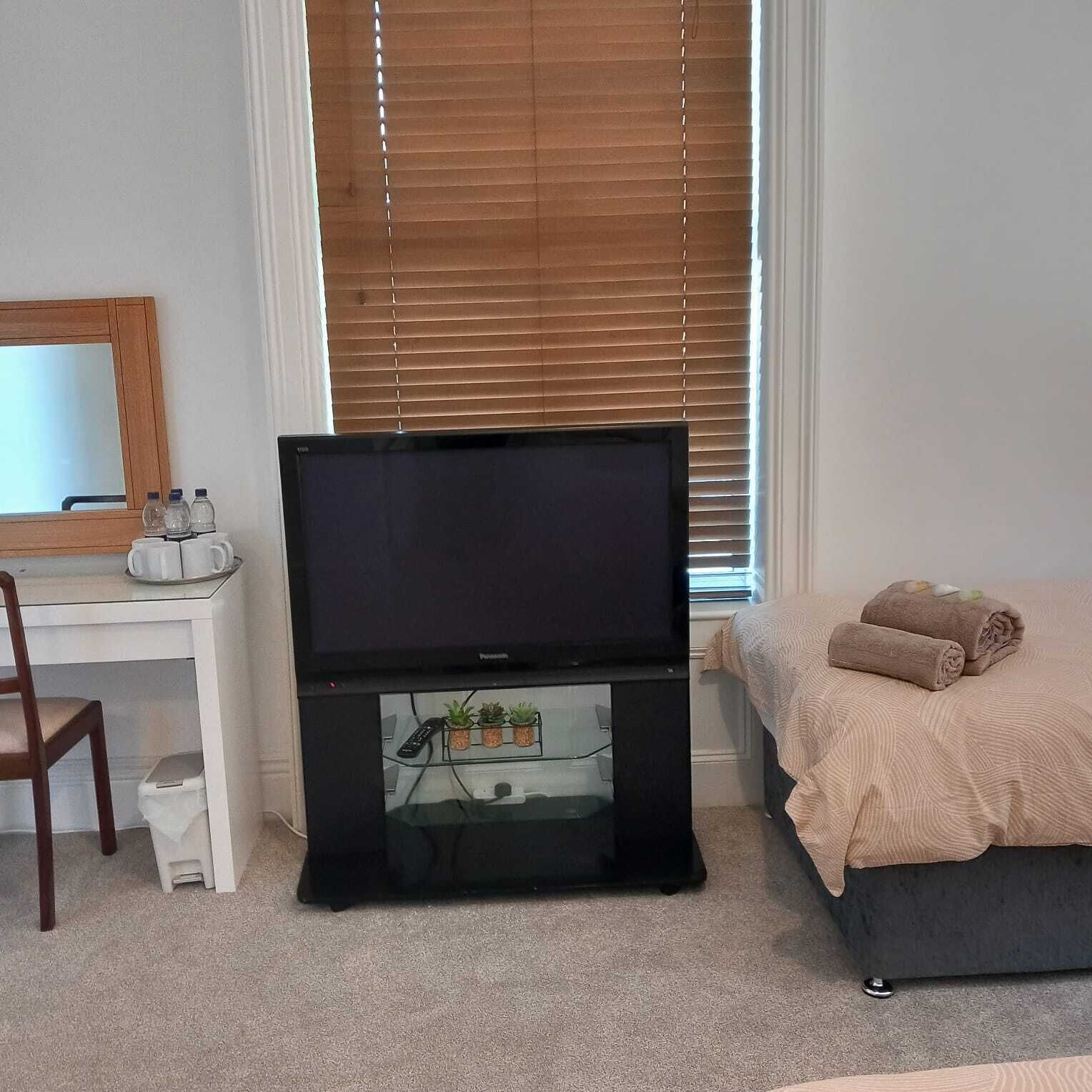 Quarto com televisão sobre suporte, escrivaninha com cadeira, cama com toalhas e janela com persianas.