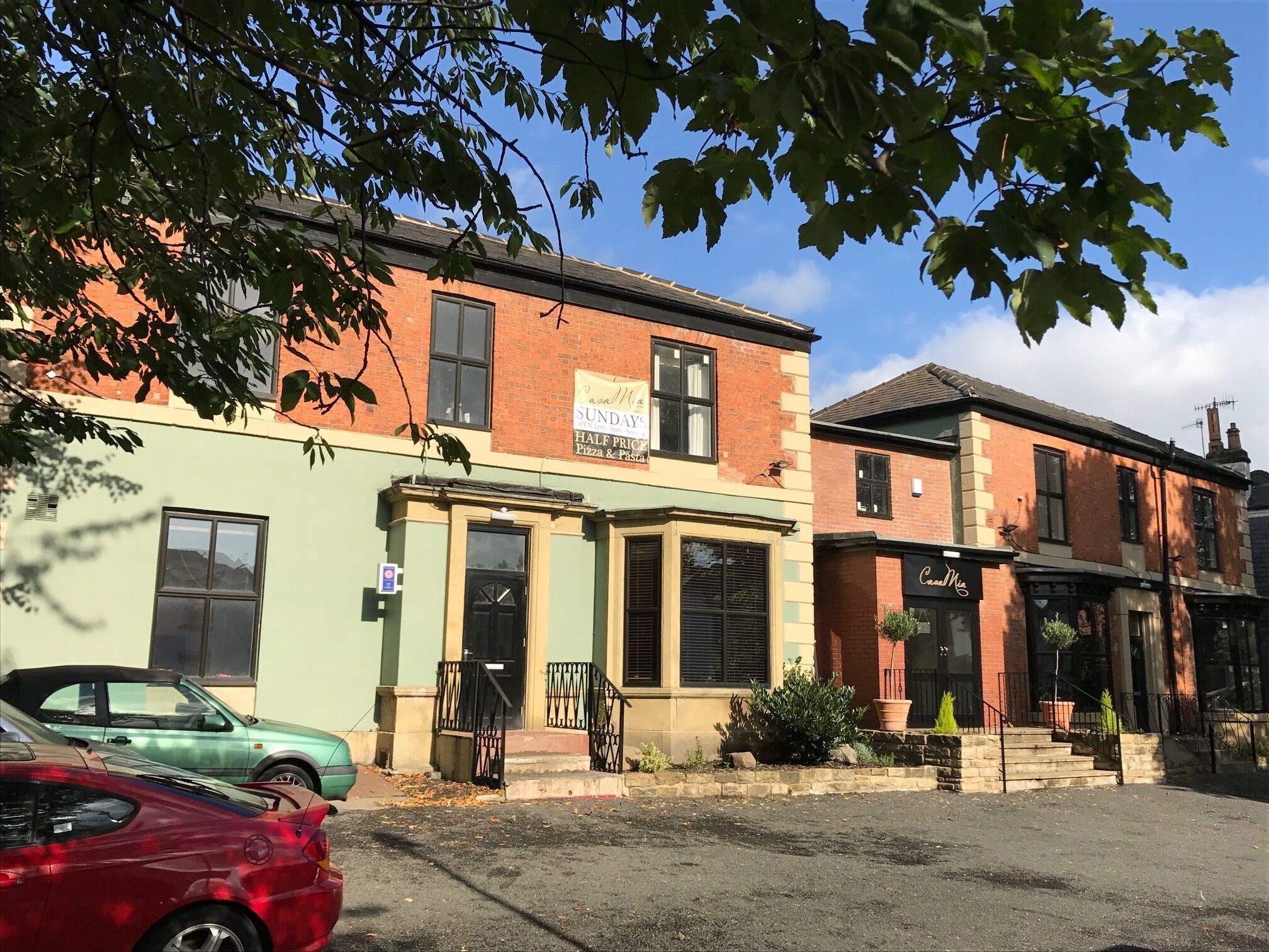 A row of buildings with a sign for 'Pasal M' and 'Sunday's Half Price Pizza & Pasta'. There are cars parked in front, and a tree with green leaves in the foreground.