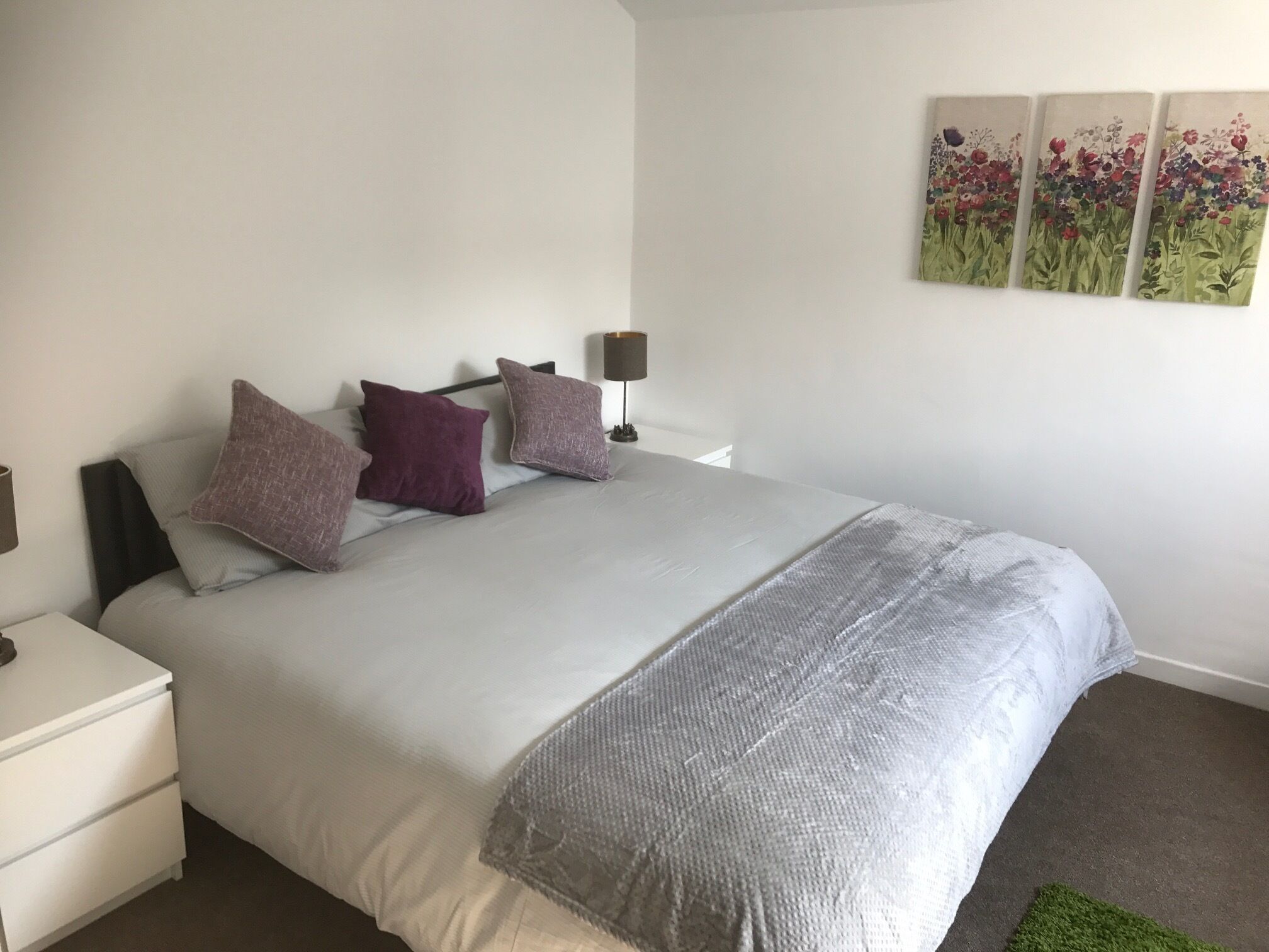 Un dormitorio con una cama, almohadas, una mesita de noche, una lámpara y una pared decorada con tres cuadros.