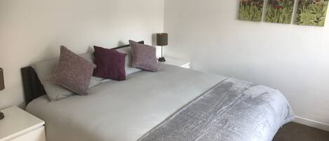 Un dormitorio con una cama, almohadas, una mesita de noche, una lámpara y una pared decorada con tres cuadros.