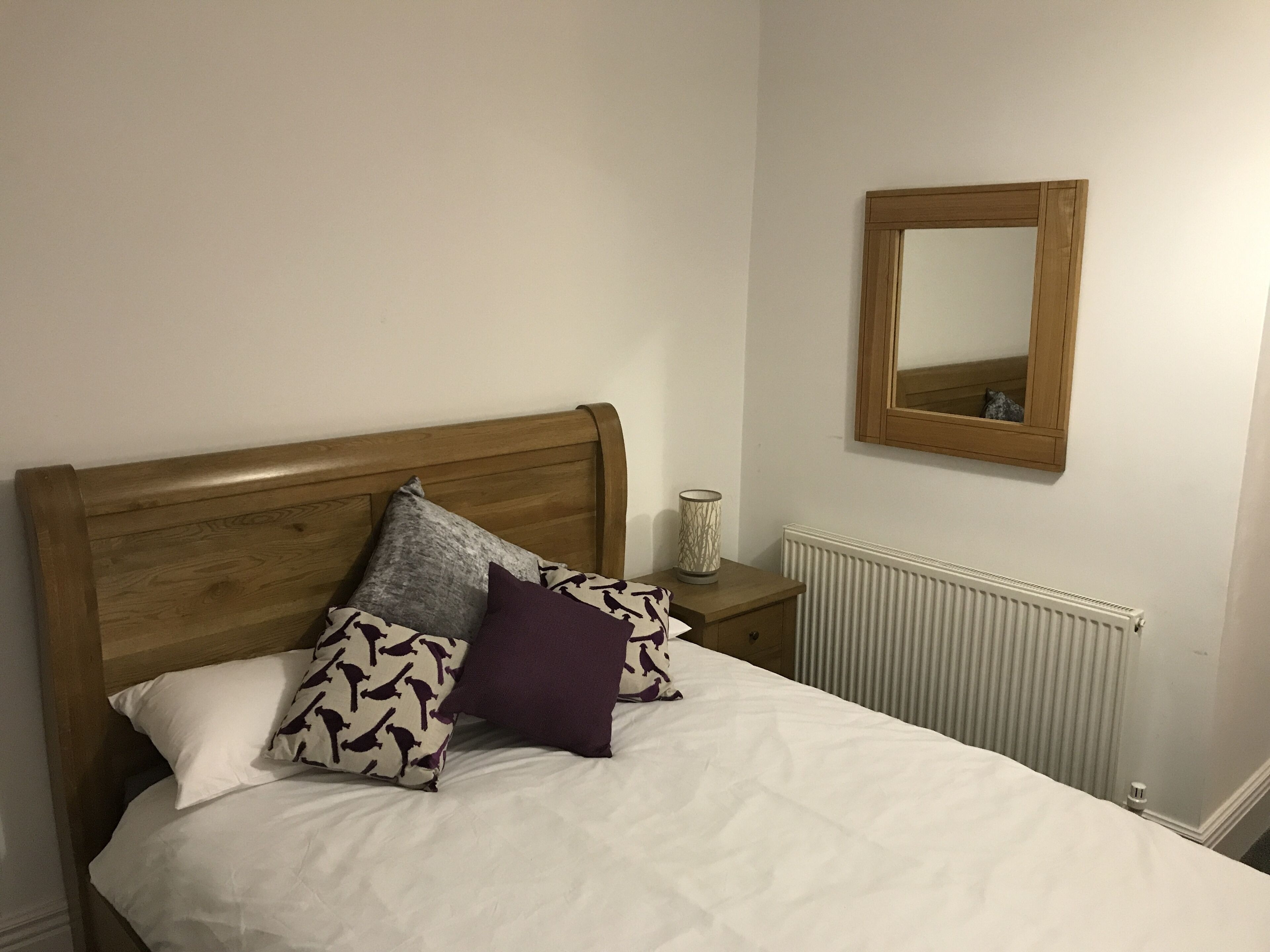 A bed with a wooden headboard, white bedding, and patterned pillows. A bedside table with a lamp and a mirror is visible.