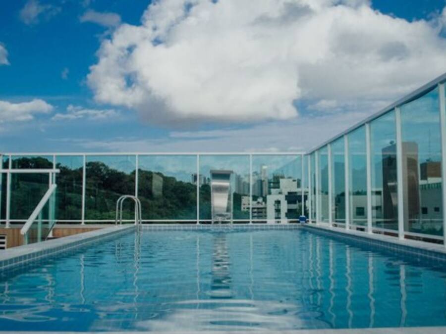 Piscina com água azul cristalina, rodeada por paredes de vidro e vista para a cidade.