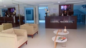 A reception area with a reception desk, two sofas, a small table with magazines, and a TV screen. - Oriental Praia Hotel (João Pessoa)