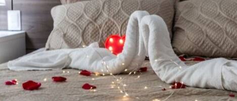 Una cama bien tendida con sábanas blancas y una decoración en forma de corazón rojo, rodeada de pétalos de rosa y luces de hadas sobre un suelo de madera.