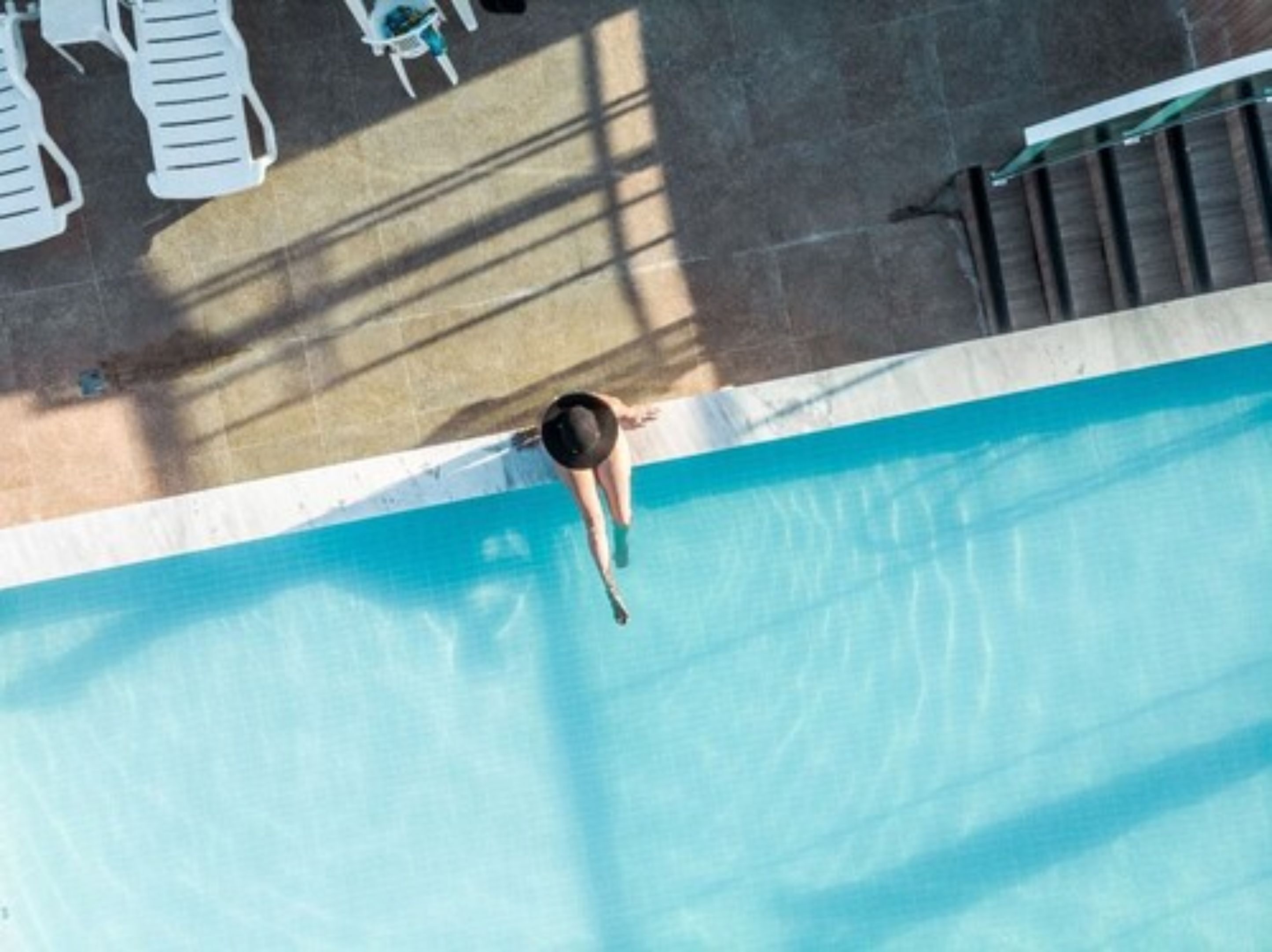 Une vue aérienne d’une piscine dans laquelle une personne plonge, entourée de chaises longues et d’un escalier.