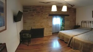 A bedroom with a stone wall, a bed with a headboard, a small table, and a window with curtains. - La Posada de Pedrazales (Galende)