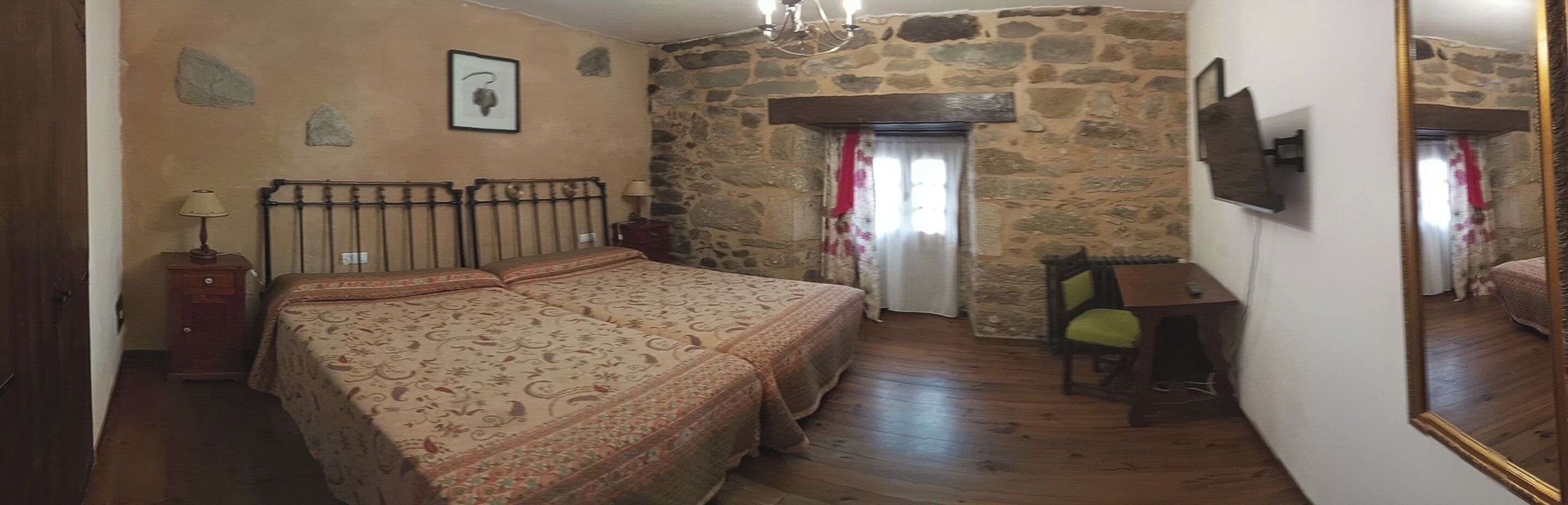 Quarto com parede de pedra, cama com colcha estampada, mesa de cabeceira, abajur, cadeira e uma pequena mesa.