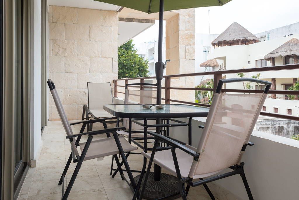 A balcony with a round table and chairs, a green umbrella, and a view of buildings with thatched roofs.