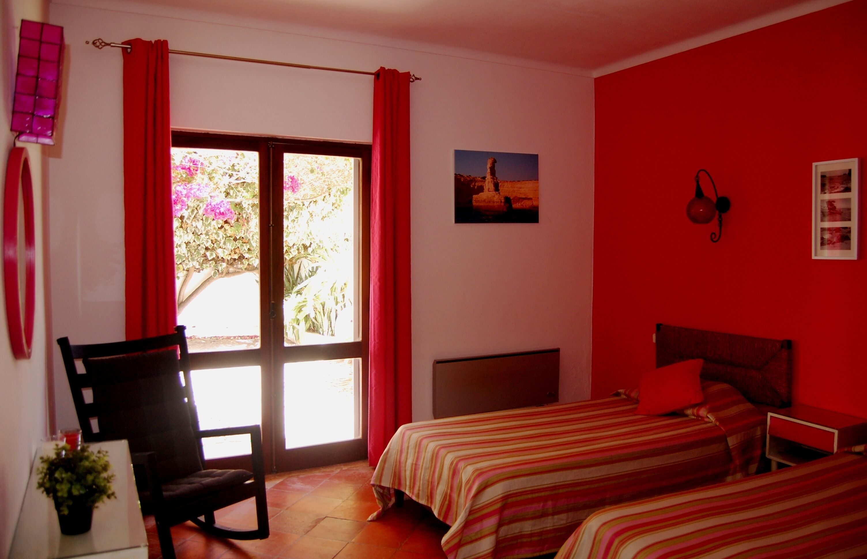 A room with two beds, a chair, a window with red curtains, a wall-mounted lamp, and a framed picture.