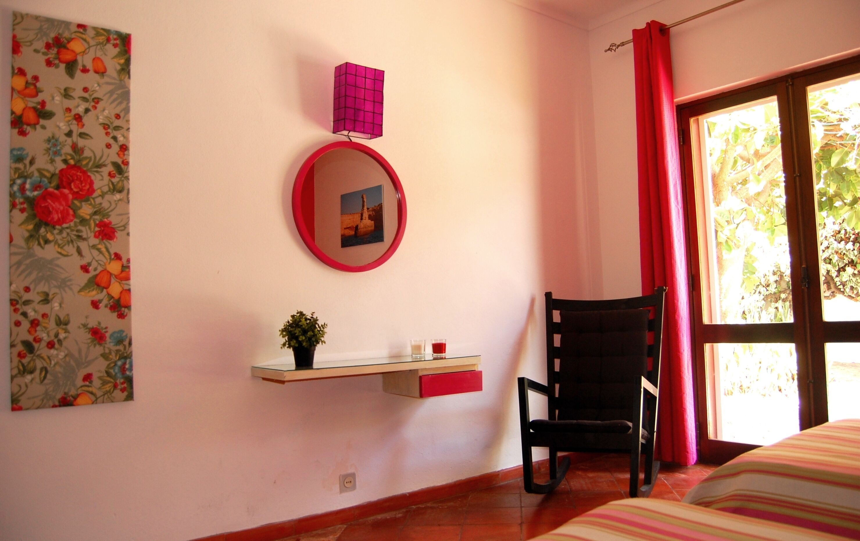 A room with a bed, a chair, a small table with a plant, a mirror, and a window with curtains.