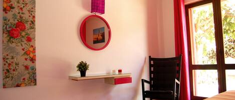 A room with a bed, a chair, a small table with a plant, a mirror, and a window with curtains.