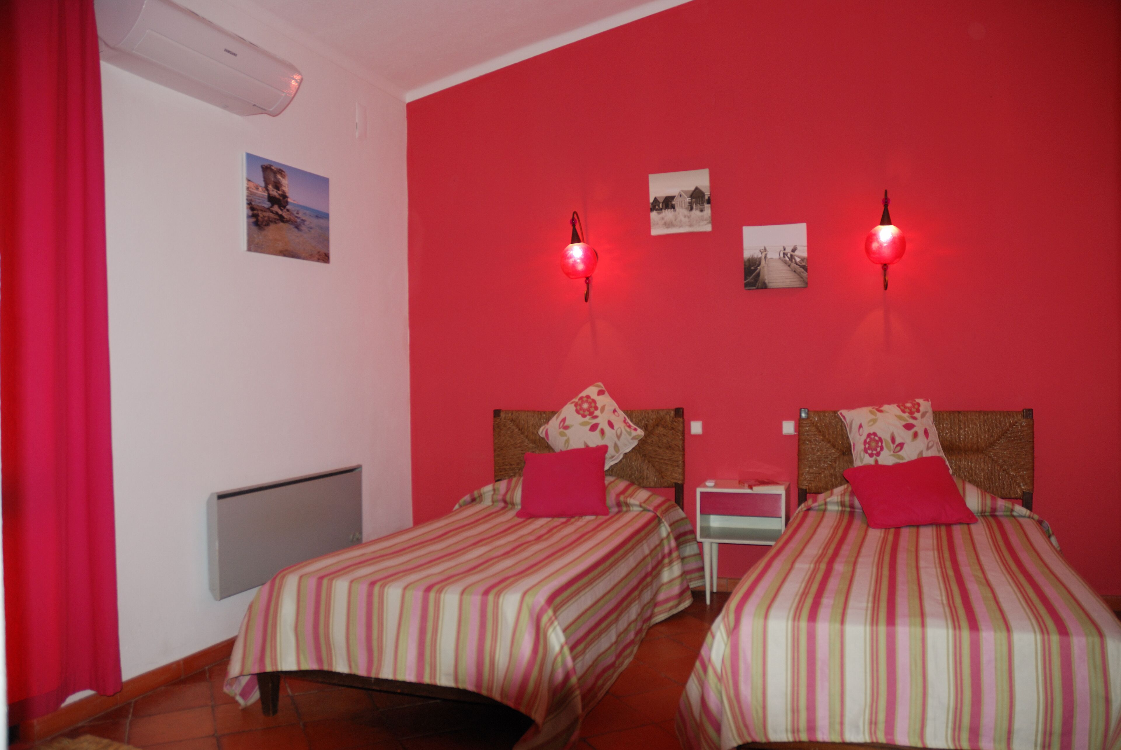 Een kamer met twee bedden, elk voorzien van gestreept beddengoed en rode kussens, een rode muur, en een kleine tafel met een lamp.