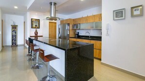 A modern kitchen with a central island, bar stools, wooden cabinets, stainless steel appliances, and a decorative mirror. - Studio one 208 by CocoBR (Playa del Carmen)
