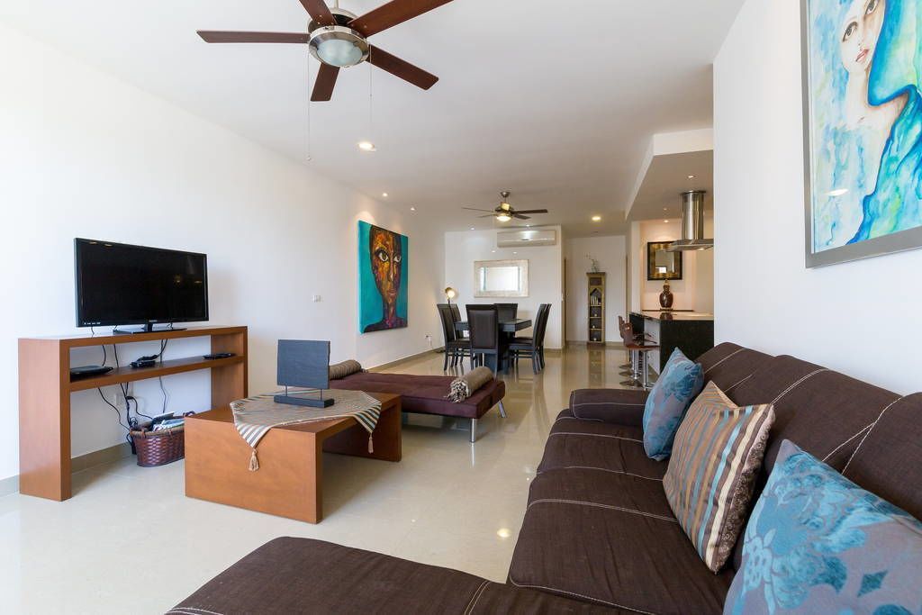Uma sala de estar moderna com uma TV de tela plana, um conjunto de sofás, uma área de jantar e uma cozinha ao fundo.