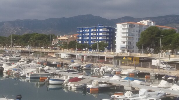 A marina with boats docked, a building with balconies, and mountains in the background.