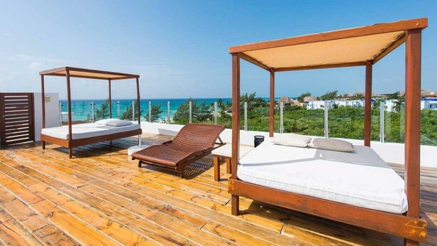 A wooden deck with two wooden beds and a lounge chair, overlooking a clear blue sea and greenery.