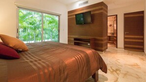 A bedroom with a large bed, a TV mounted on a wooden wall, and a sliding glass door leading to a balcony with greenery.
