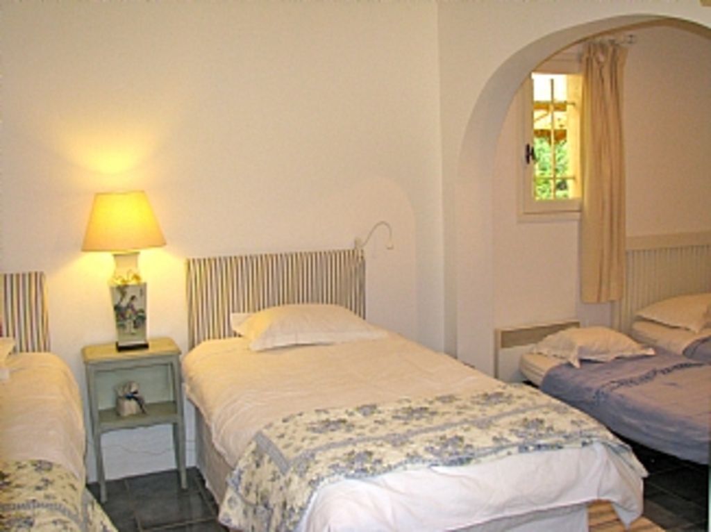 A bedroom with two beds, a bedside table with a lamp, a small shelf, and a window with curtains.