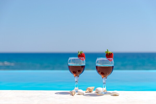 Two glasses of red wine with strawberries on a white surface by a pool with a clear blue sky.