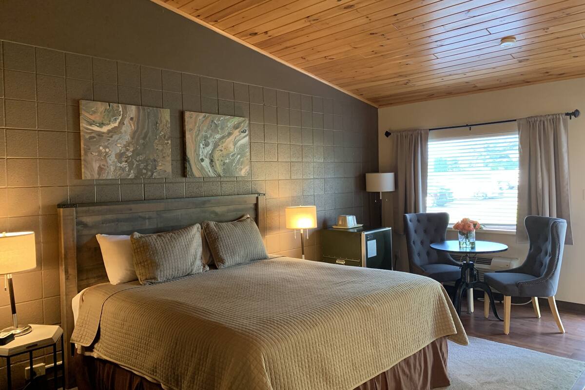 A hotel room with a large bed, a wooden ceiling, a gray tiled wall, and a window with curtains.
