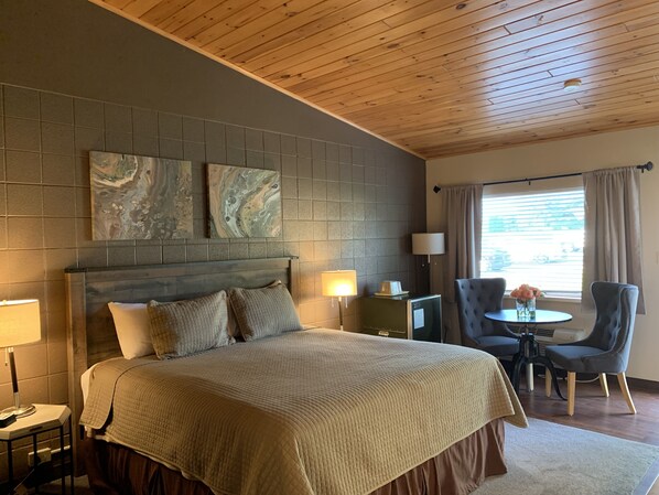 Habitación de hotel con una cama grande, techo de madera, pared con azulejos grises y una ventana con cortinas.