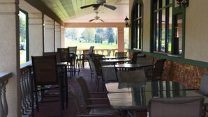 A covered outdoor dining area with tables and chairs, large windows, and a view of a park.