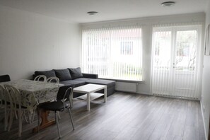 Un salon moderne comprenant un canapé, une table basse, une table à manger et des chaises. De grandes fenêtres équipées de stores, un radiateur et une porte donnant sur l’extérieur complètent l’ensemble.
