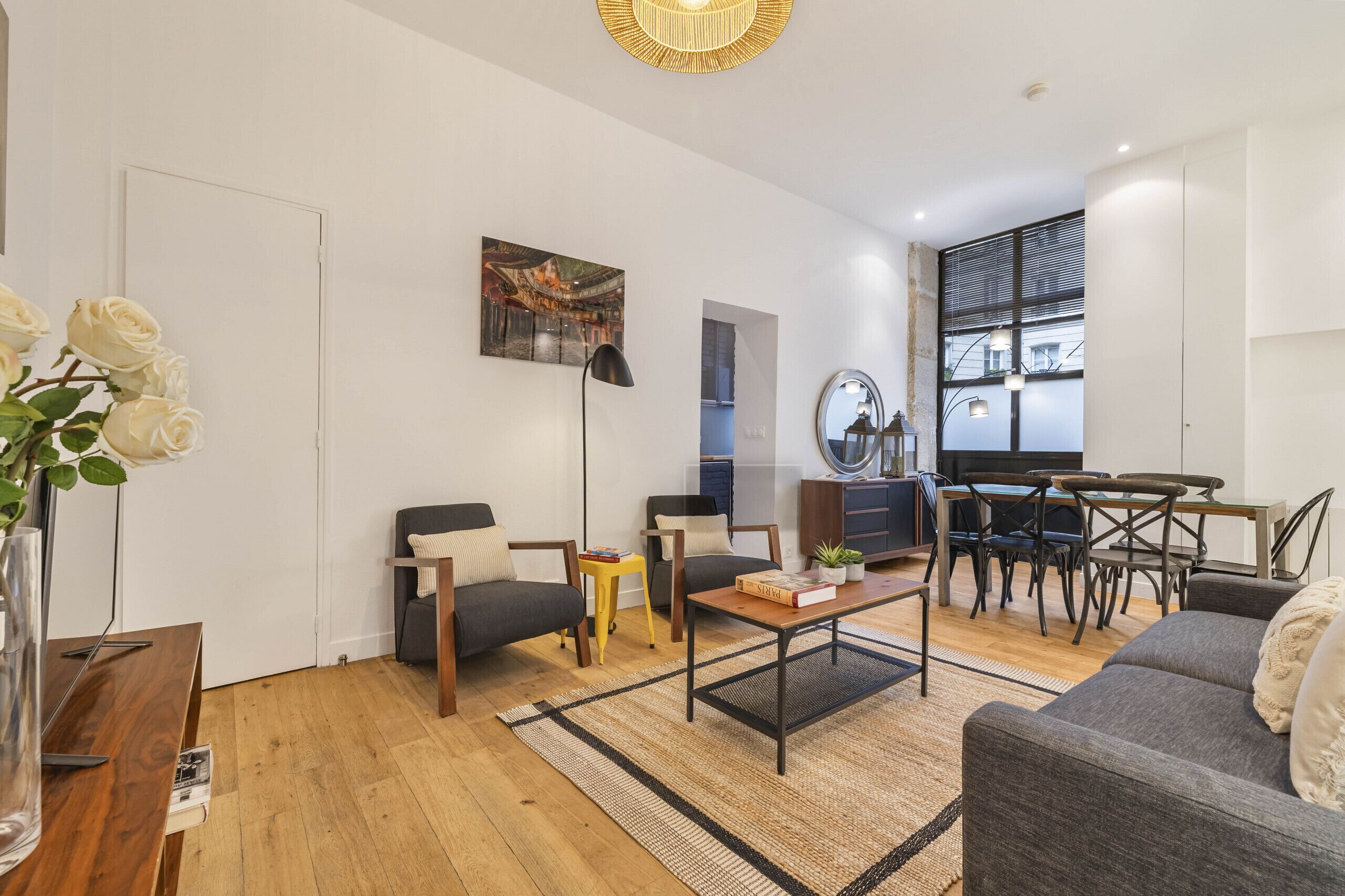 A modern living room with a sofa, coffee table, and chairs, a dining area with a table and chairs, and a large window with blinds.