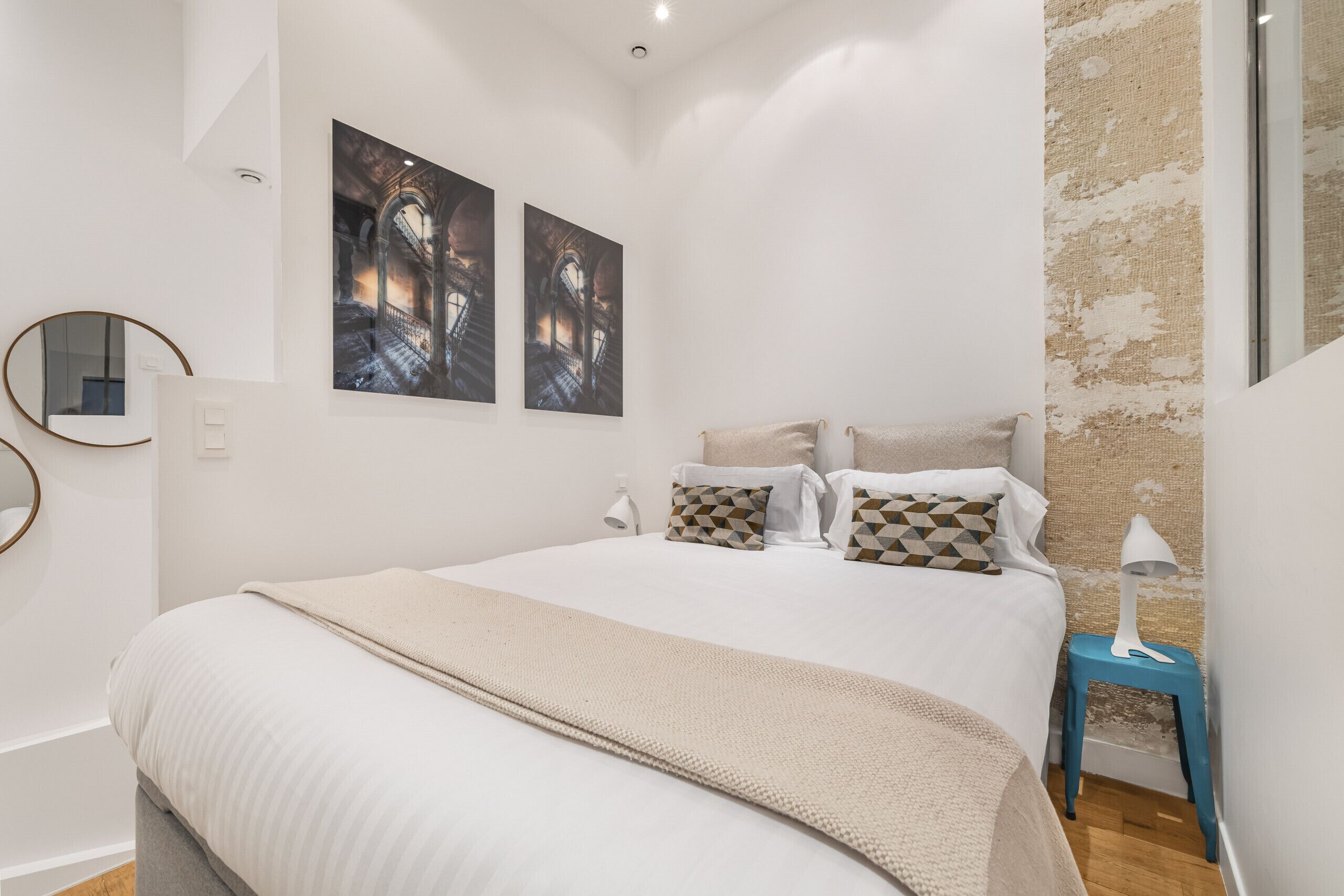 Une chambre à coucher avec un grand lit, deux œuvres d’art accrochées au mur, un miroir rond et une petite table de chevet avec une lampe.