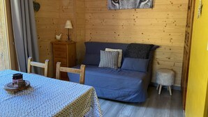 Een gezellige kamer met houten inrichting, een blauwe bank met kussens, een klein tafeltje met een tafelkleed met dessin, en een aan de muur hangend schilderij.