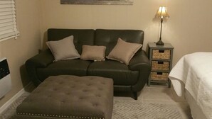 Une pièce avec un canapé marron foncé, un pouf assorti, une table de chevet avec une lampe et un tableau représentant une scène de plage accroché au mur.