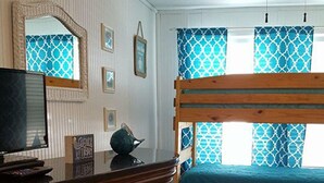 Ein Zimmer mit einem Holzkoffer, einem Stockbett mit blauer Bettwäsche, einem Deckenventilator und einem Fenster mit gemusterten Vorhängen.