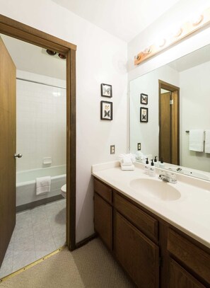 Une salle de bain avec une baignoire, des toilettes et un grand miroir. Des tableaux encadrés ornent le mur et une serviette est posée sur le sol.