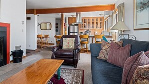 A living room with a dark brown leather armchair, a blue sofa with patterned cushions, a wooden coffee table, and a kitchen area with wooden cabinets and a refrigerator. - Outpost: Sleeping Indian E12 - Air Conditioning! (Teton Village)