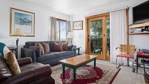 A living room with a sofa, a wooden coffee table, a red patterned rug, a television, and a sliding glass door leading to an outdoor area. - Outpost: Sleeping Indian E12 - Air Conditioning! (Teton Village)
