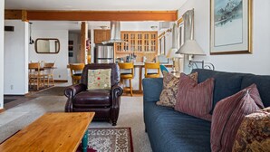 A living room with a brown leather sofa, a blue couch, and a wooden coffee table. There is a kitchen in the background with wooden cabinets and a stainless steel range hood. - Outpost: Sleeping Indian E12 - Air Conditioning! (Teton Village)