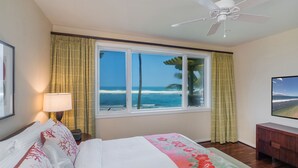 Habitación de hotel con un ventanal que ofrece vistas a la playa y al océano, una cama con ropa de cama floral, una mesita de noche con lámpara, y un televisor sobre un mueble de madera.