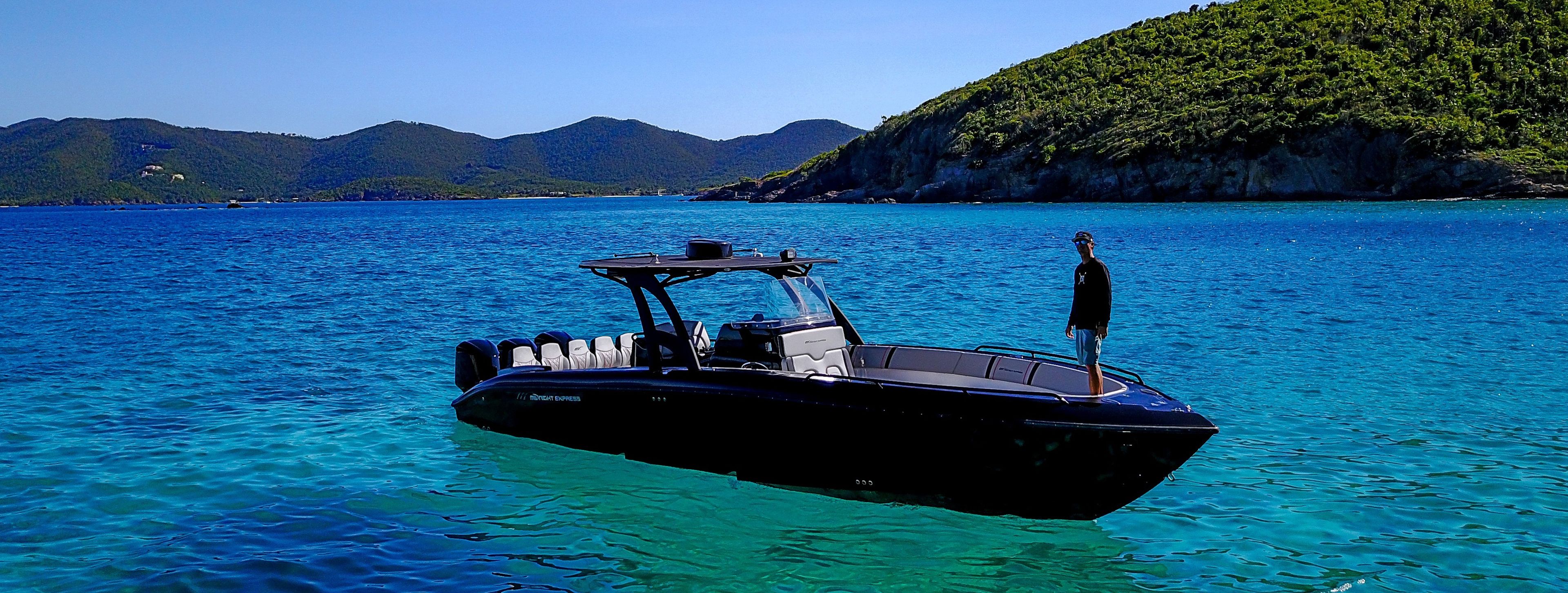 A boat with a person standing on the deck, surrounded by clear blue water and a mountainous landscape in the background.