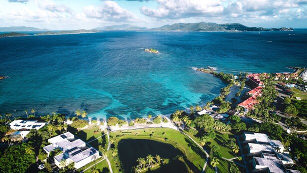 An aerial view of a coastal area with clear blue waters, a small island, and a resort with buildings and palm trees. - Beachfront Villas at Crystal Cove (St. Thomas)