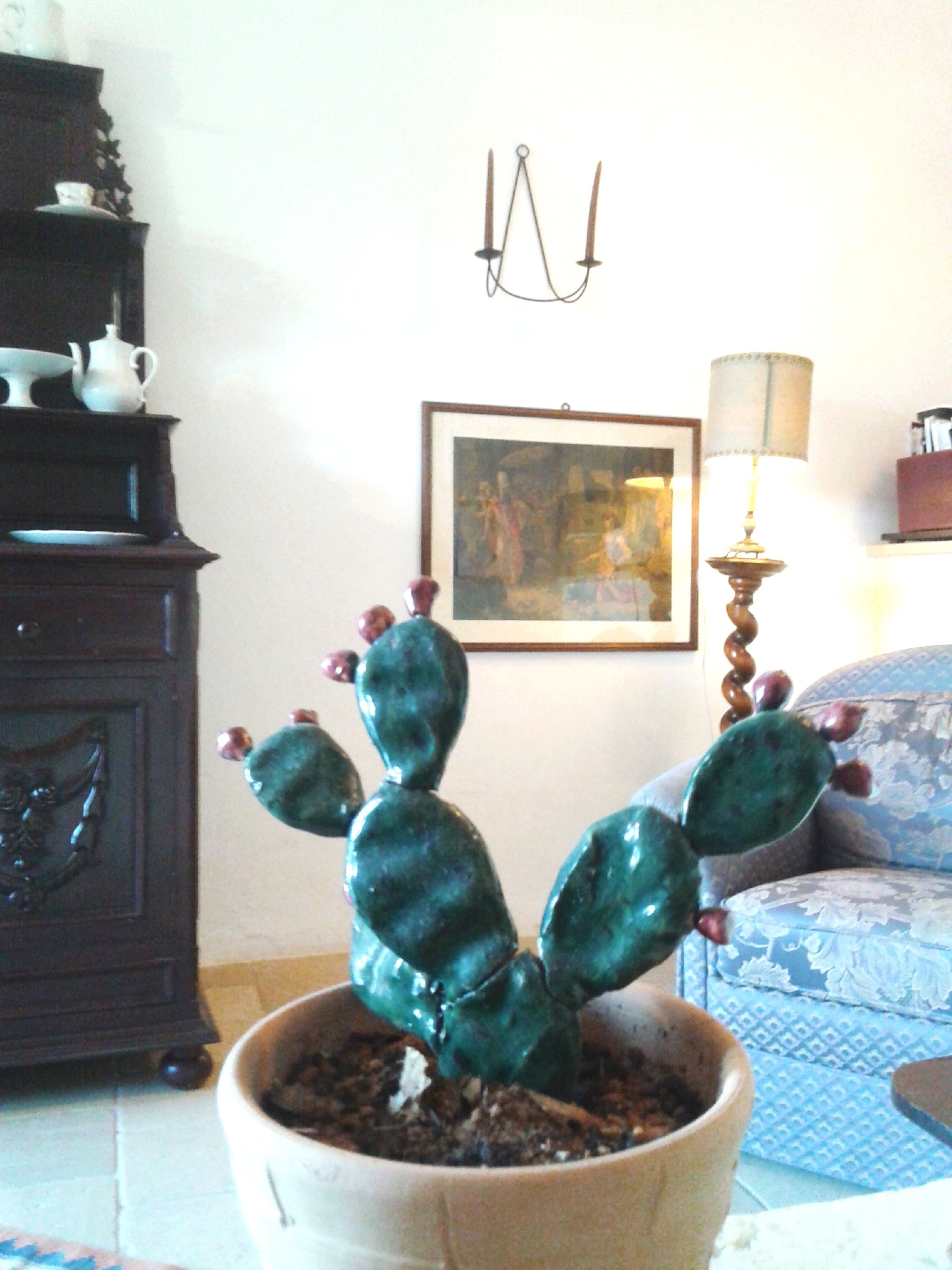 A cactus plant in a pot on a tiled floor, a dark wooden cabinet with decorative items, a framed painting, and a lamp on a table.