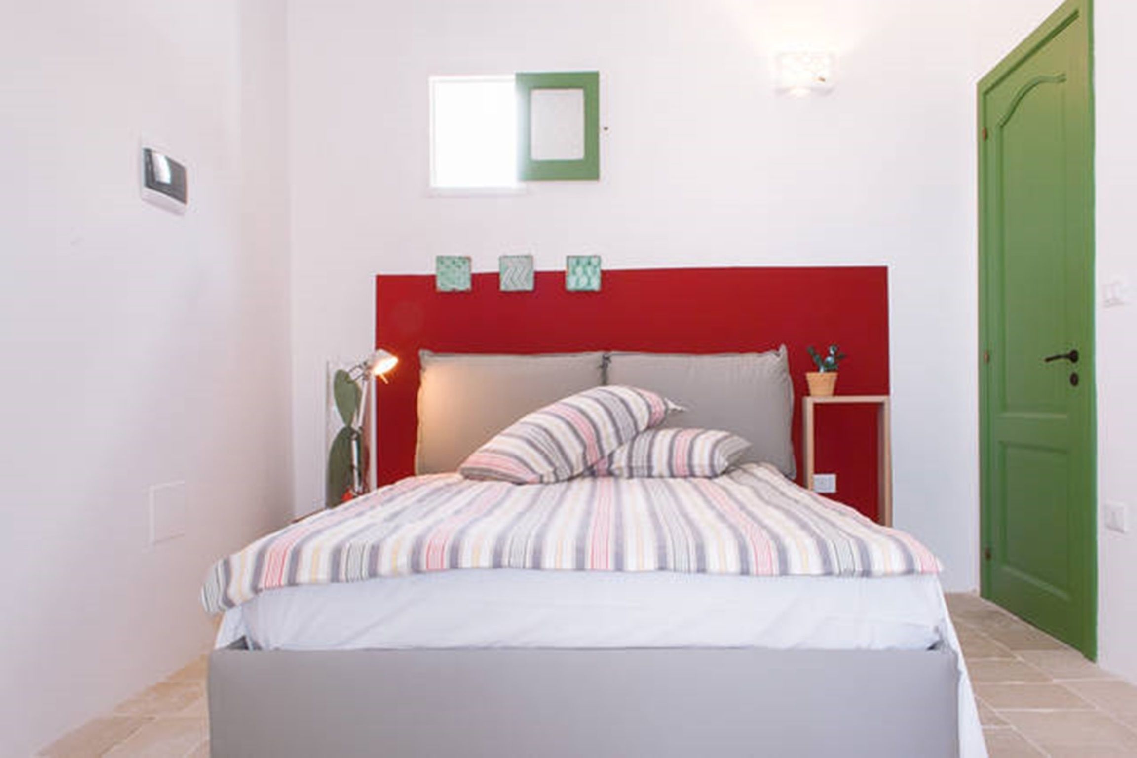 Un dormitorio con cama, cabecera roja, mesita de noche con lámpara y puerta verde.
