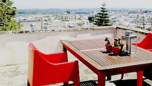 Terraza en la azotea con una mesa de madera y sillas rojas, con vista a un paisaje urbano con edificios y un árbol.